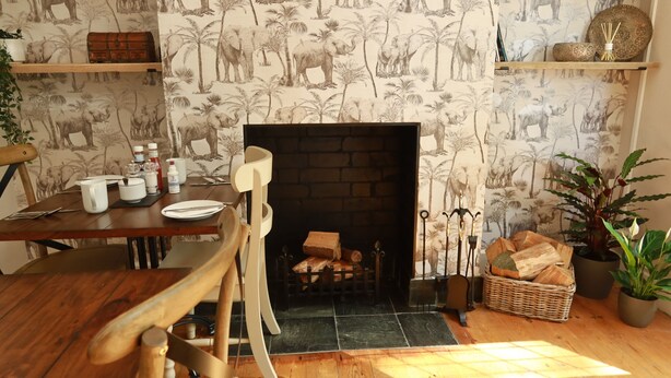 A cozy living room with a fireplace, wooden furniture, and a patterned wallpaper featuring elephants and palm trees. - Avonlea Guest House (Banbury)