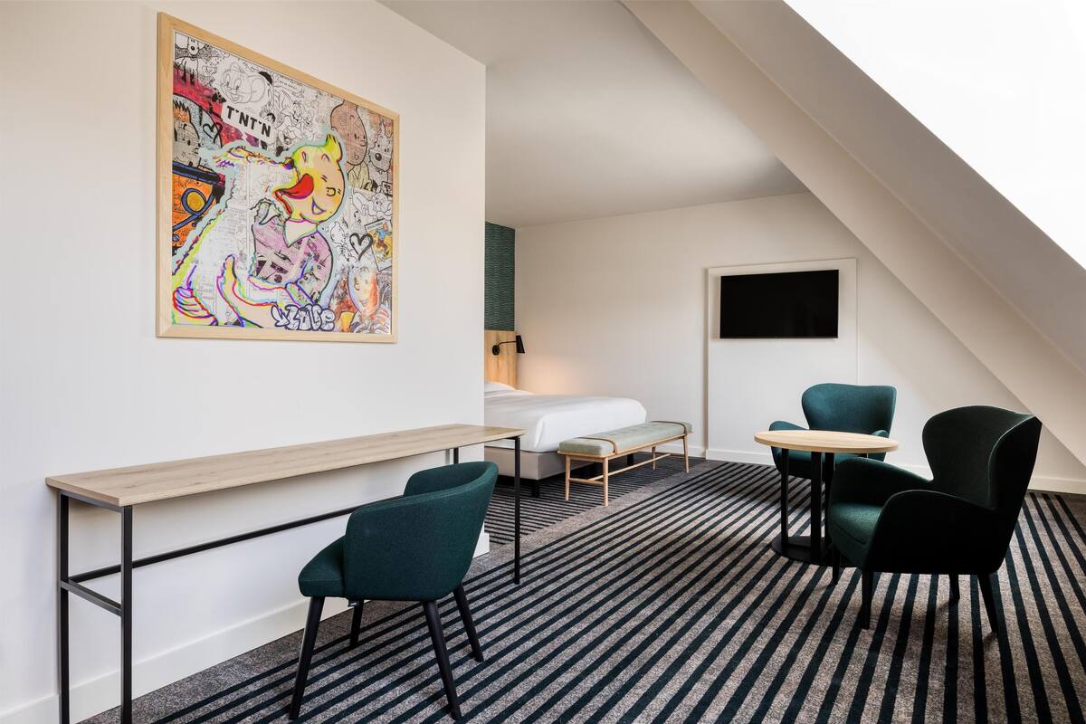 A modern hotel room with a bed, a desk, two chairs, a large abstract painting, and a TV.