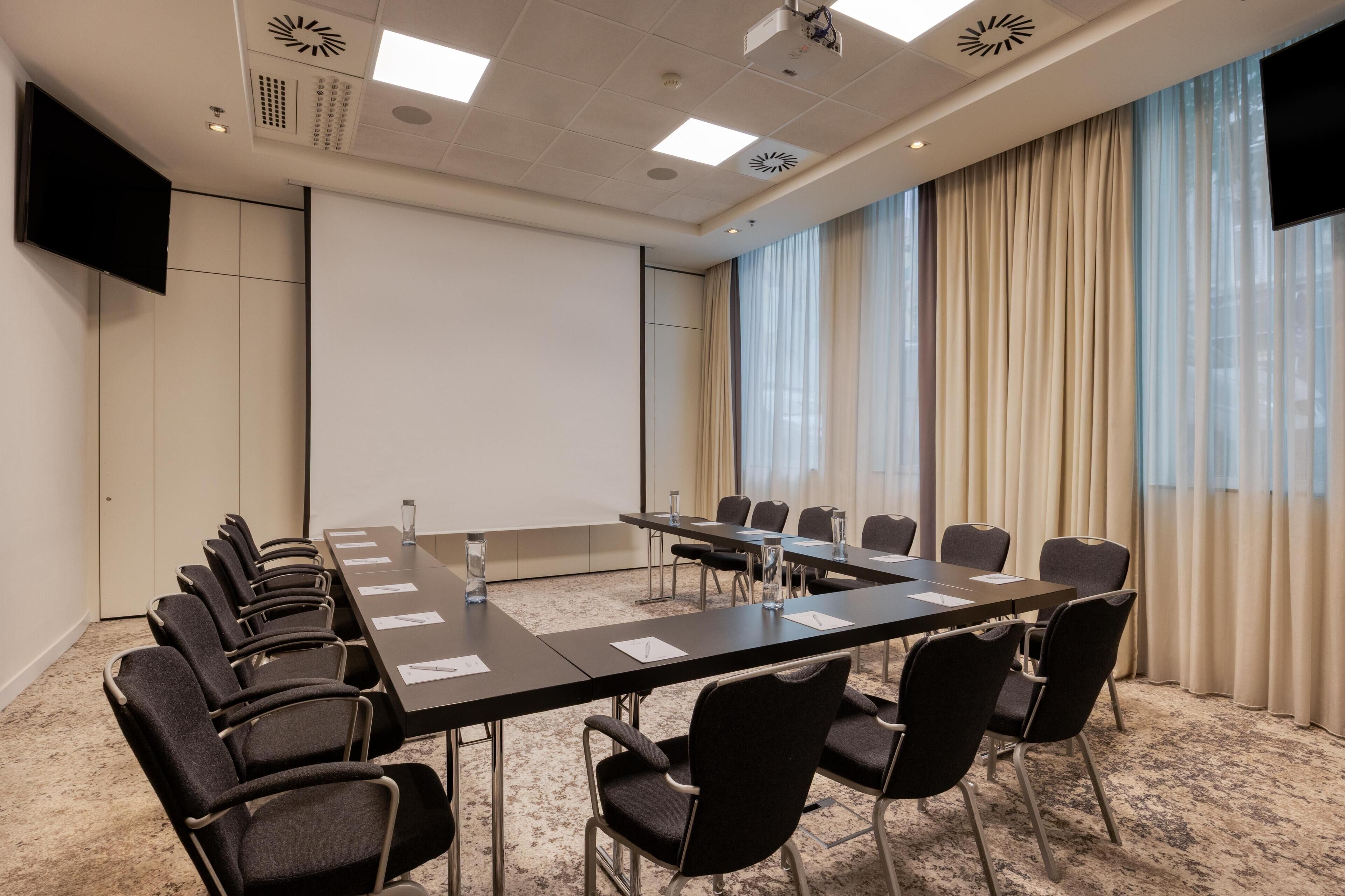 Une salle de conférence équipée d’une longue table, de chaises, d’un écran de projection et d’une grande fenêtre avec des rideaux.