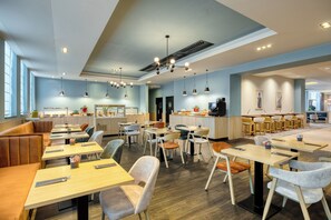 A modern dining area with wooden tables and chairs, a kitchen in the background, and a large window providing natural light. - Radisson Hotel Brussels Centre Midi (Brussels)