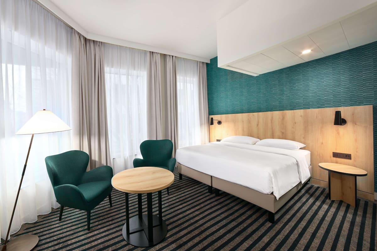 A hotel room with a bed, two chairs, a small table, a lamp, and a large window with curtains.