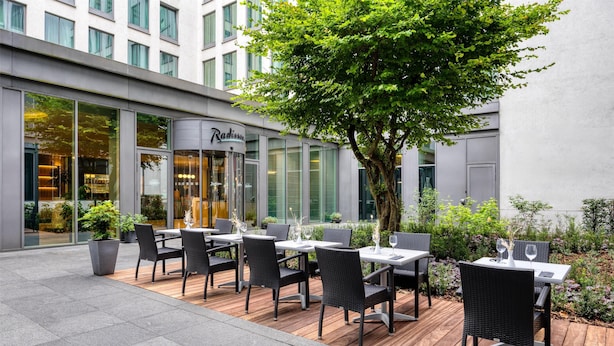 Un área de comedor al aire libre con mesas y sillas, un árbol grande y un edificio con ventanas amplias y un cartel que dice "Radisson".