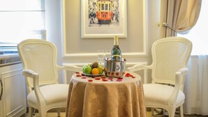 Una mesa de comedor con una botella de champán, fruta y copas, rodeada de sillas blancas en una habitación con un cuadro en la pared.