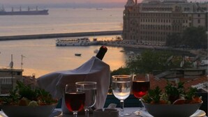 Ein gedeckter Tisch mit Weingläsern und einer Schüssel Essen, mit Blick auf ein Gewässer, im Hintergrund ein großes Schiff und ein Gebäude.