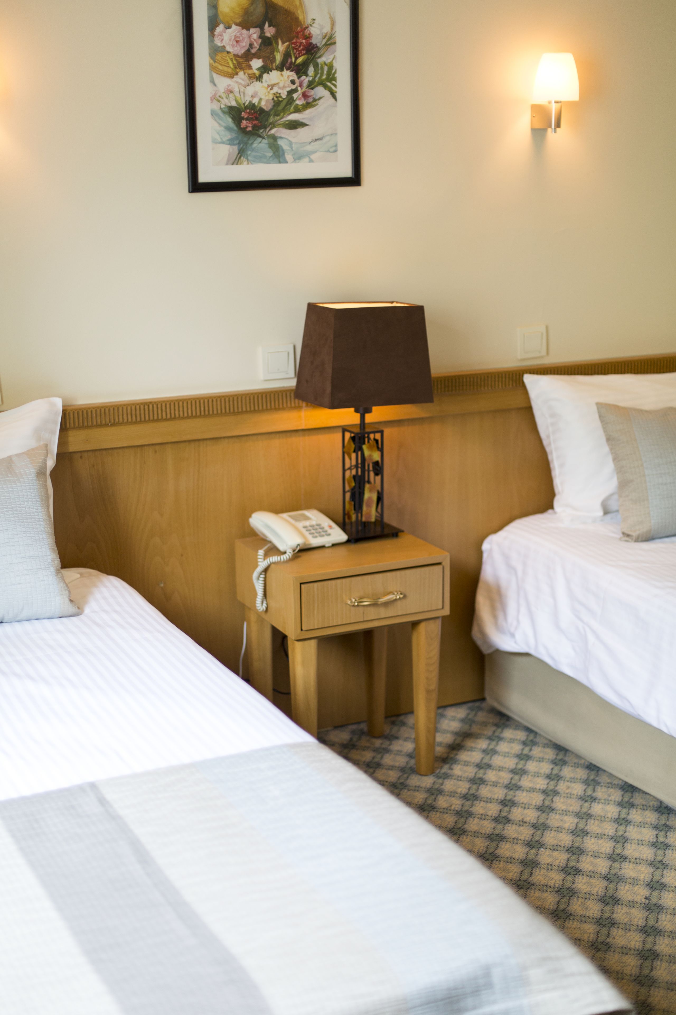 A hotel room with two beds, a bedside table with a lamp, a phone, and a wall-mounted light.