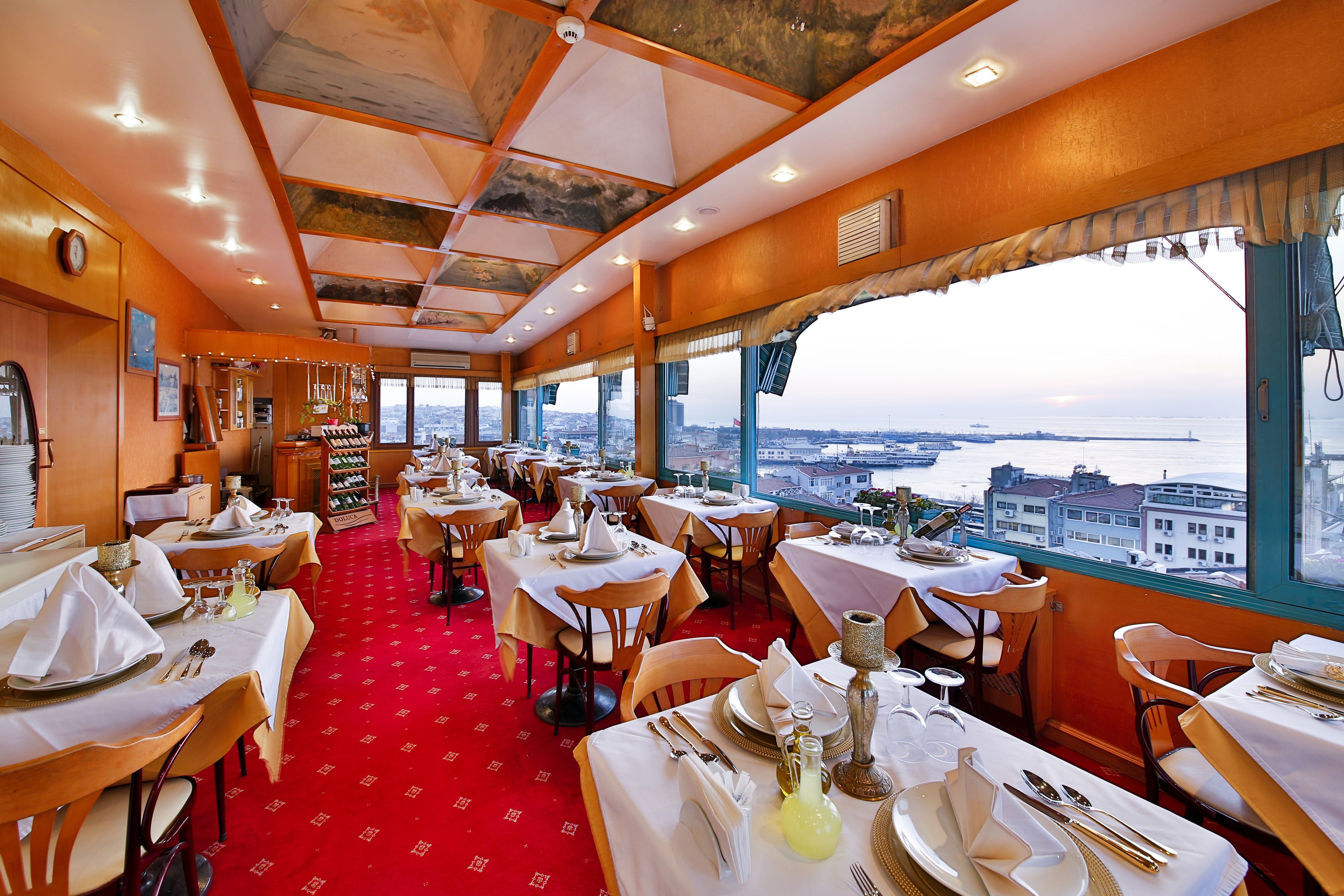 Un restaurant avec des tables dressées pour le dîner, un tapis rouge et une vue sur la ville grâce à de grandes fenêtres.