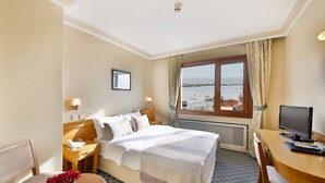 Ein Hotelzimmer mit Bett, Schreibtisch, Stuhl, Fernseher, Fenster mit Meerblick und einer Blumenvase auf dem Schreibtisch.
