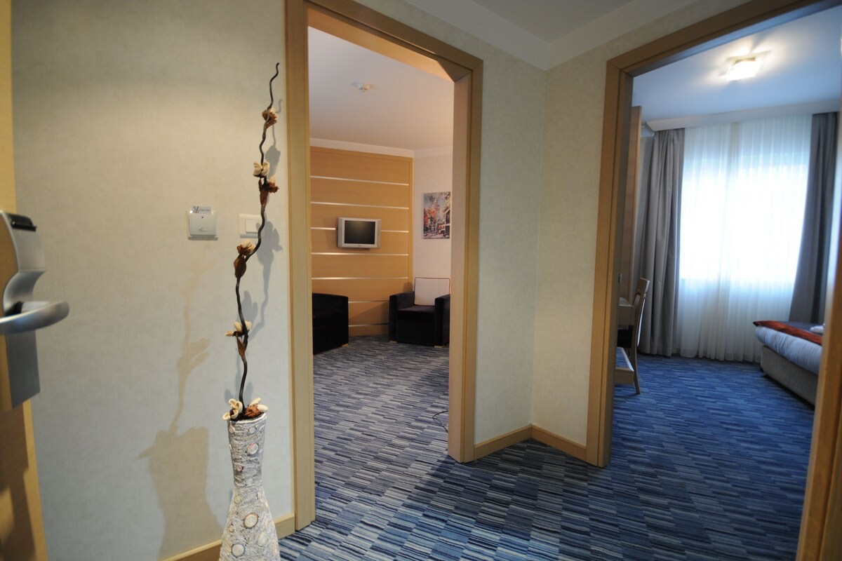 Ein Hotelzimmer mit modernem Design, inklusive Flachbildfernseher, einer Sitzecke mit Sofa und Sesseln sowie einem großen Fenster mit Vorhängen.