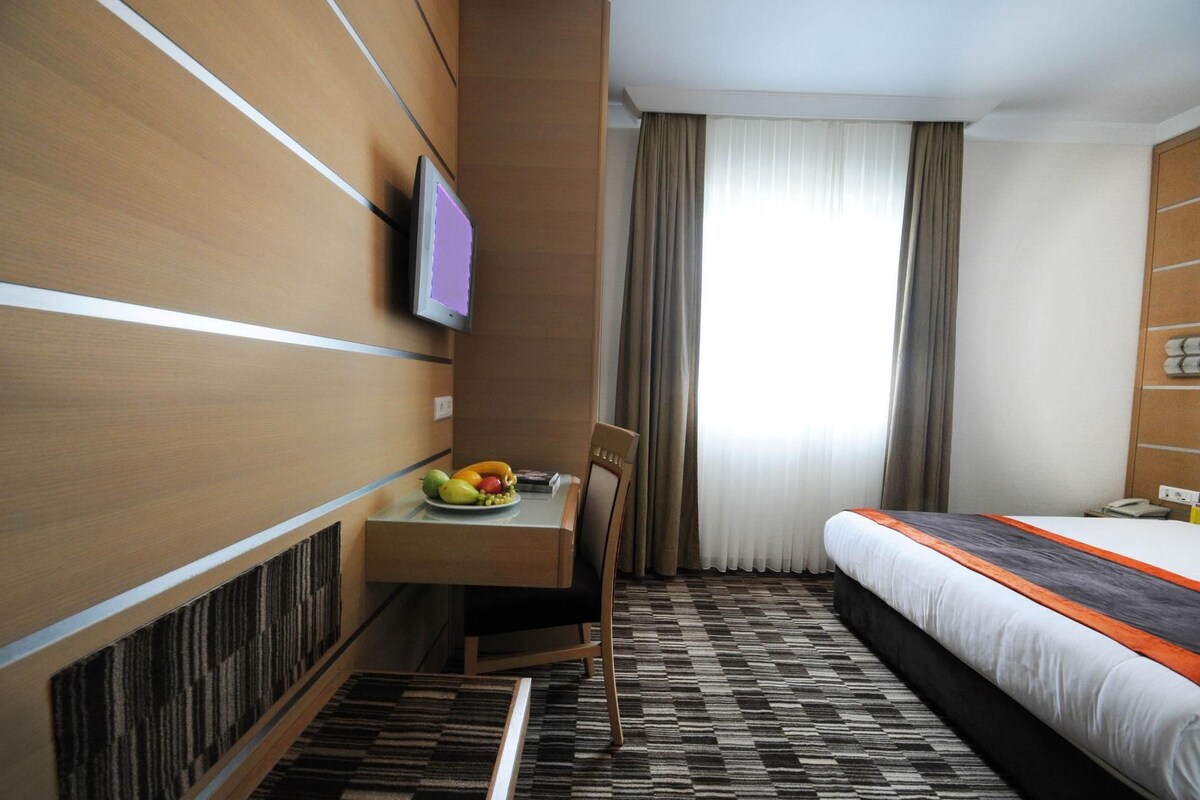 Ein Hotelzimmer mit Bett, Schreibtisch mit Obstschale, Fernseher und einem Vorhangfenster.
