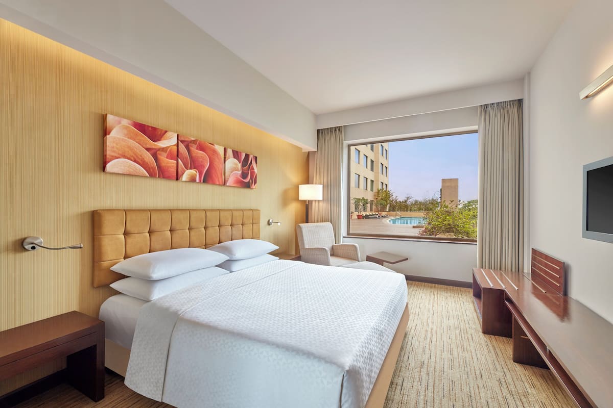 Habitación de hotel con una cama grande, un escritorio, una silla, un televisor y una ventana con vistas a edificios y una piscina.