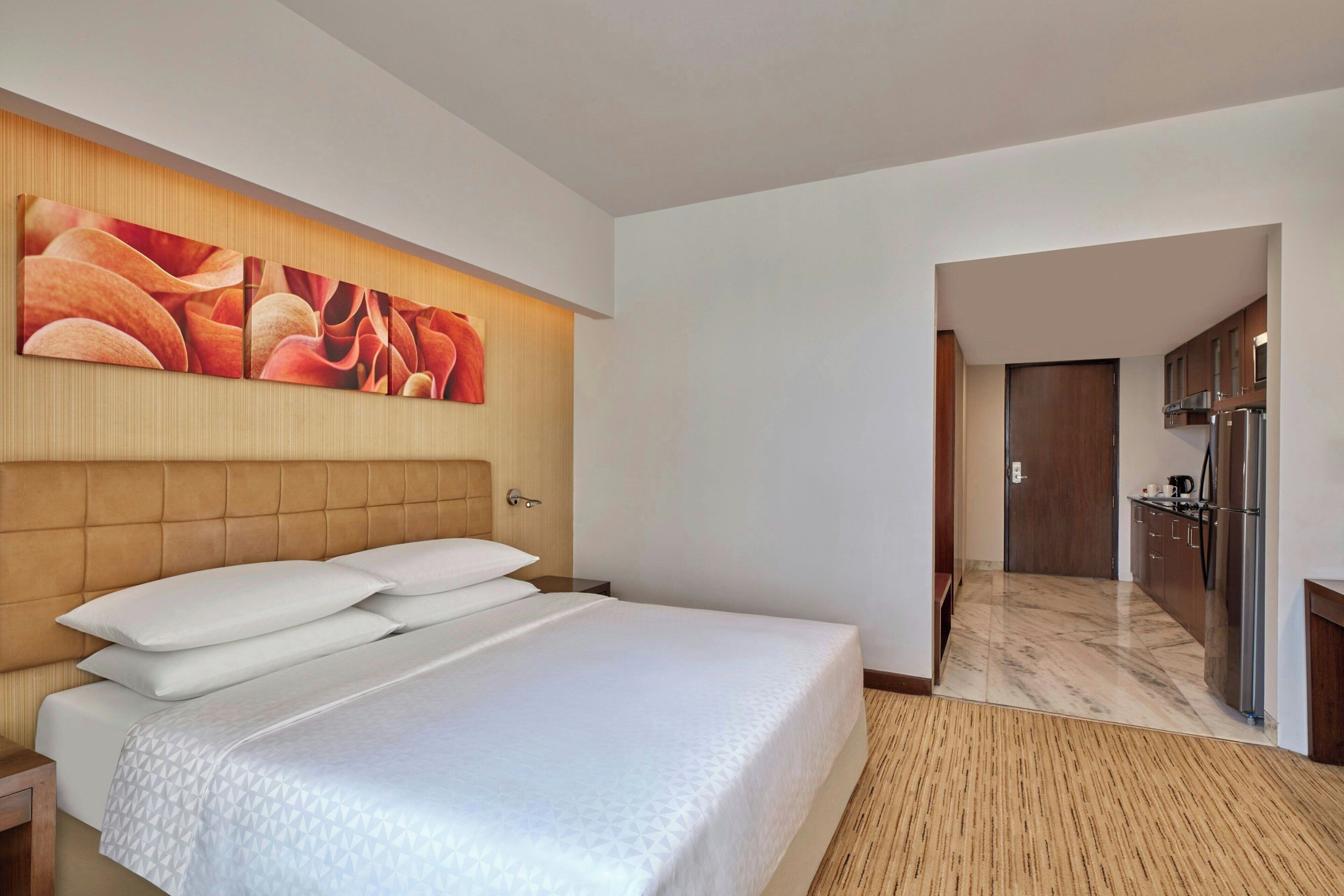 Habitació d'hotel amb un llit gran, un panell de fusta al capçal, una obra d'art penjada a la paret i vista cap a la zona de cuina.