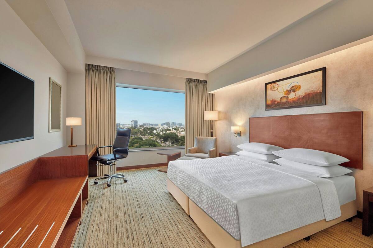 Habitación de hotel con una cama grande, un escritorio, una silla, un televisor y vista a la ciudad desde la ventana.
