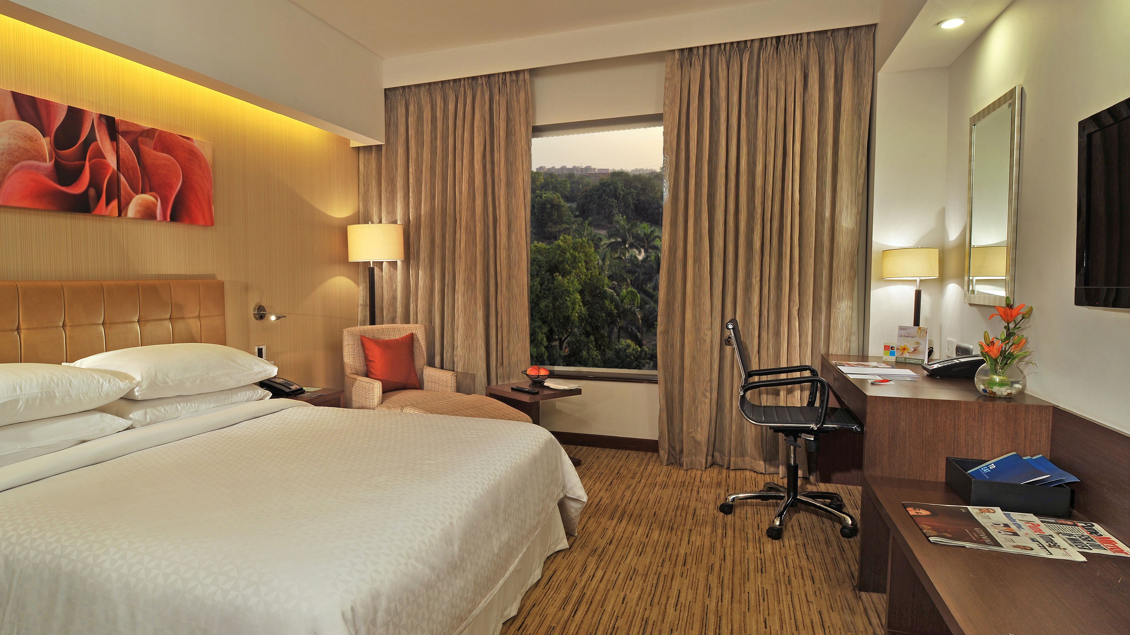 Une chambre d’hôtel avec un grand lit, un bureau avec une chaise, une lampe et une vue sur les arbres par la fenêtre.