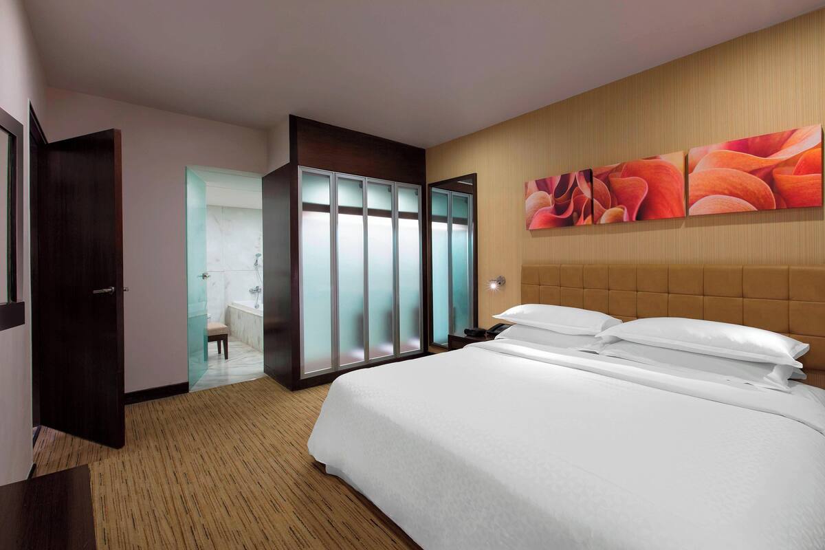 Habitación de hotel con una cama grande, un armario, un baño con puerta de vidrio y una pared con arte abstracto.