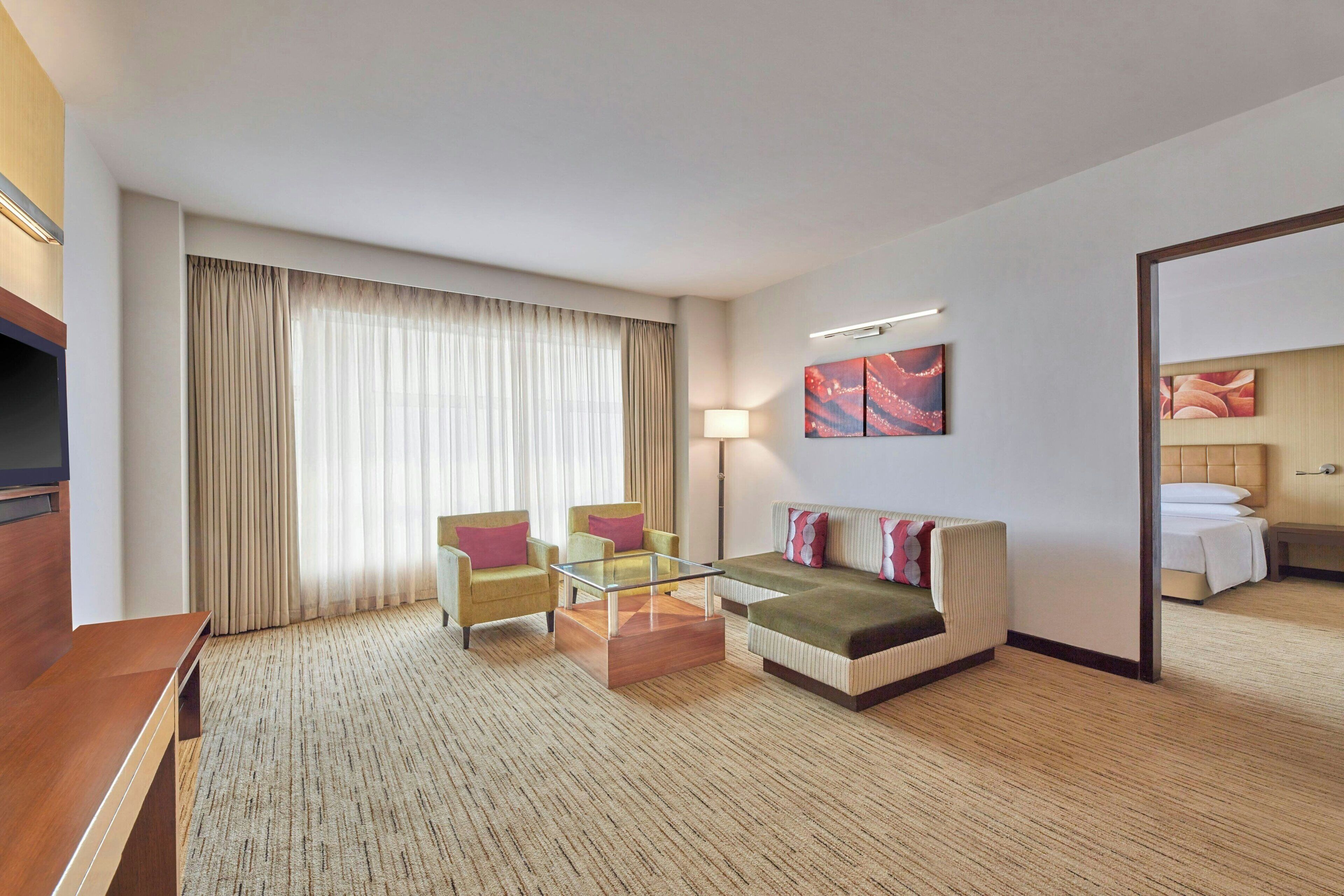A hotel room with a sofa, two armchairs, a glass coffee table, a TV, a lamp, and a painting on the wall.