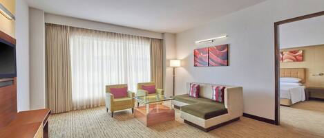 Ein Hotelzimmer mit einer Sitzecke, zwei Sesseln, einem Glastisch, einem Fernseher, einer Lampe und einem Bild an der Wand.