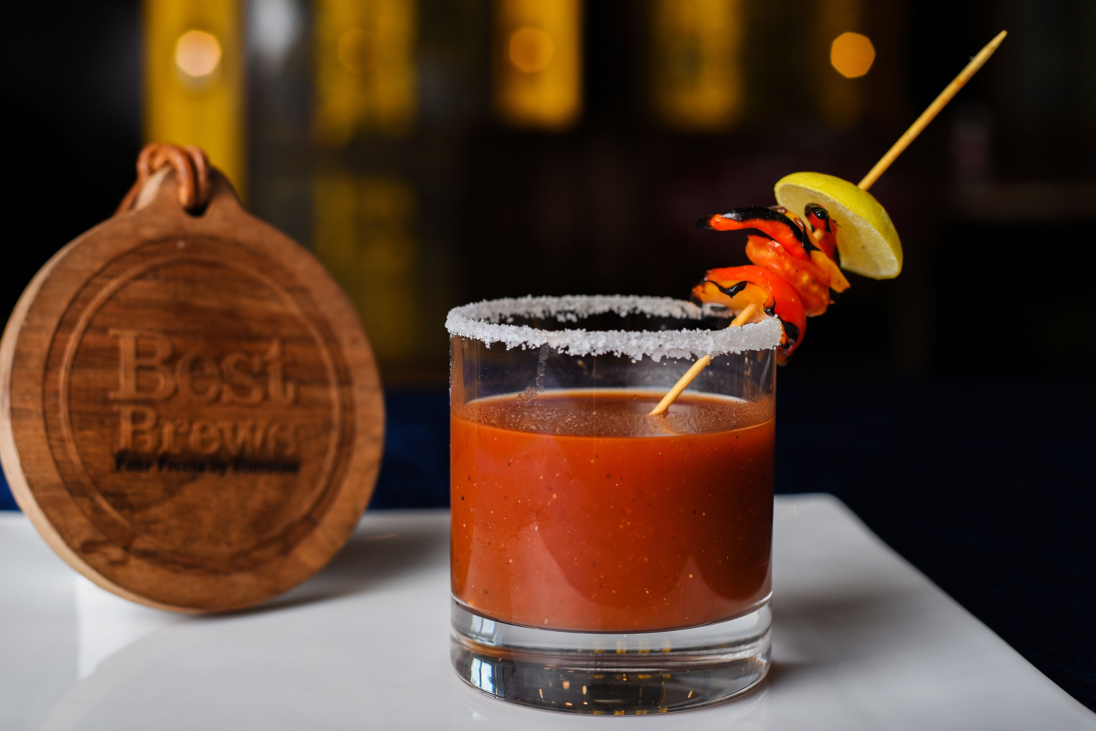 Bloody Mary in een glas, gegarneerd met een citroenpartje, op een houten onderzetter met reliëftekst, met een vage achtergrond van een bar.