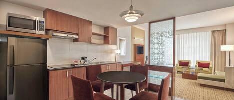 A modern kitchen with a microwave, refrigerator, and dining area with chairs and a table. There is a sofa and a lamp in the living area.