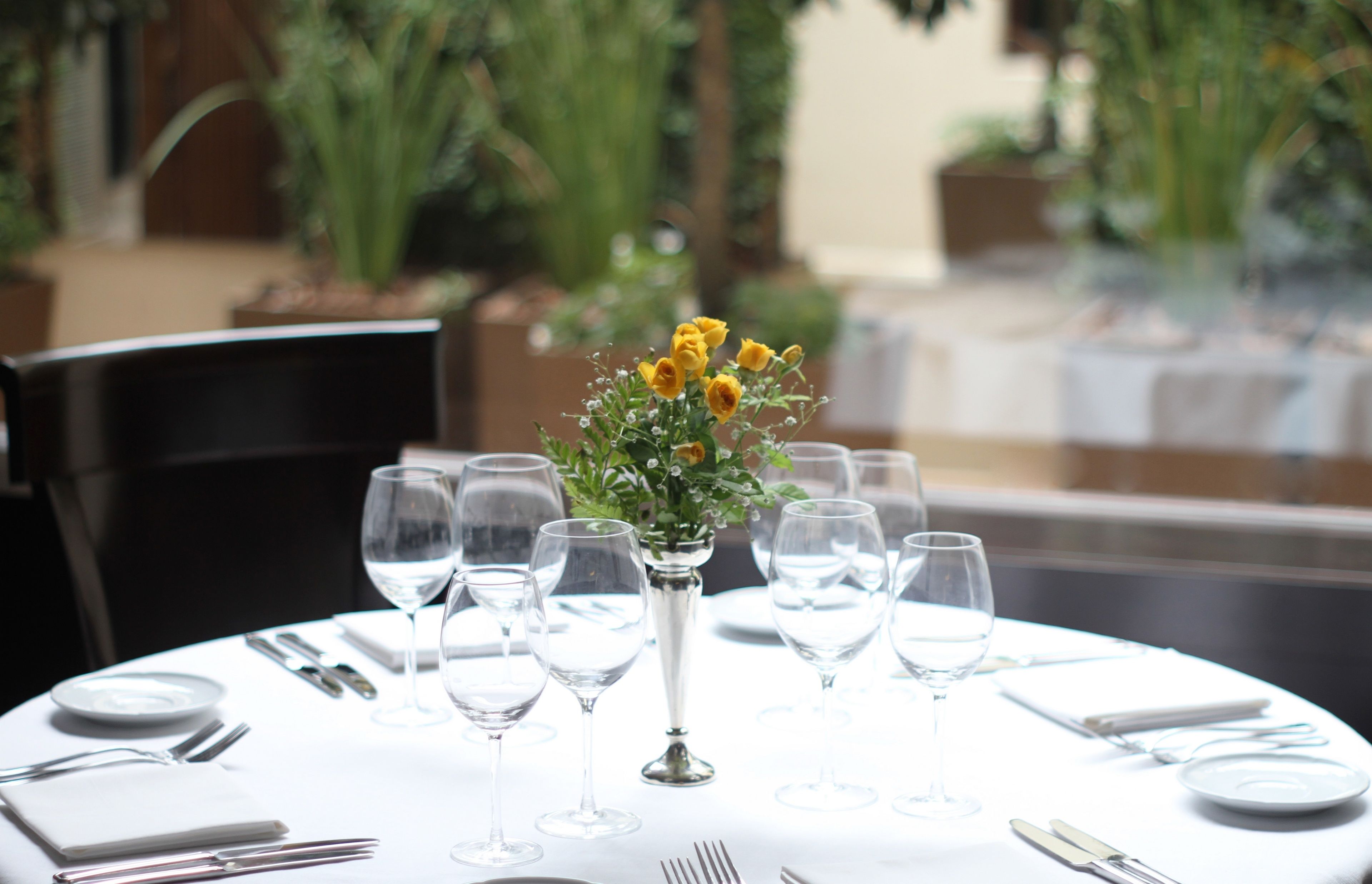 Una mesa redonda de comedor con mantel blanco, platos, cubiertos y copas de vino, adornada con un jarrón de flores amarillas en el centro.