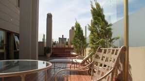 Un balcón con mesa de vidrio y sillas de mimbre, con vista a un paisaje urbano con edificios y cielo despejado.