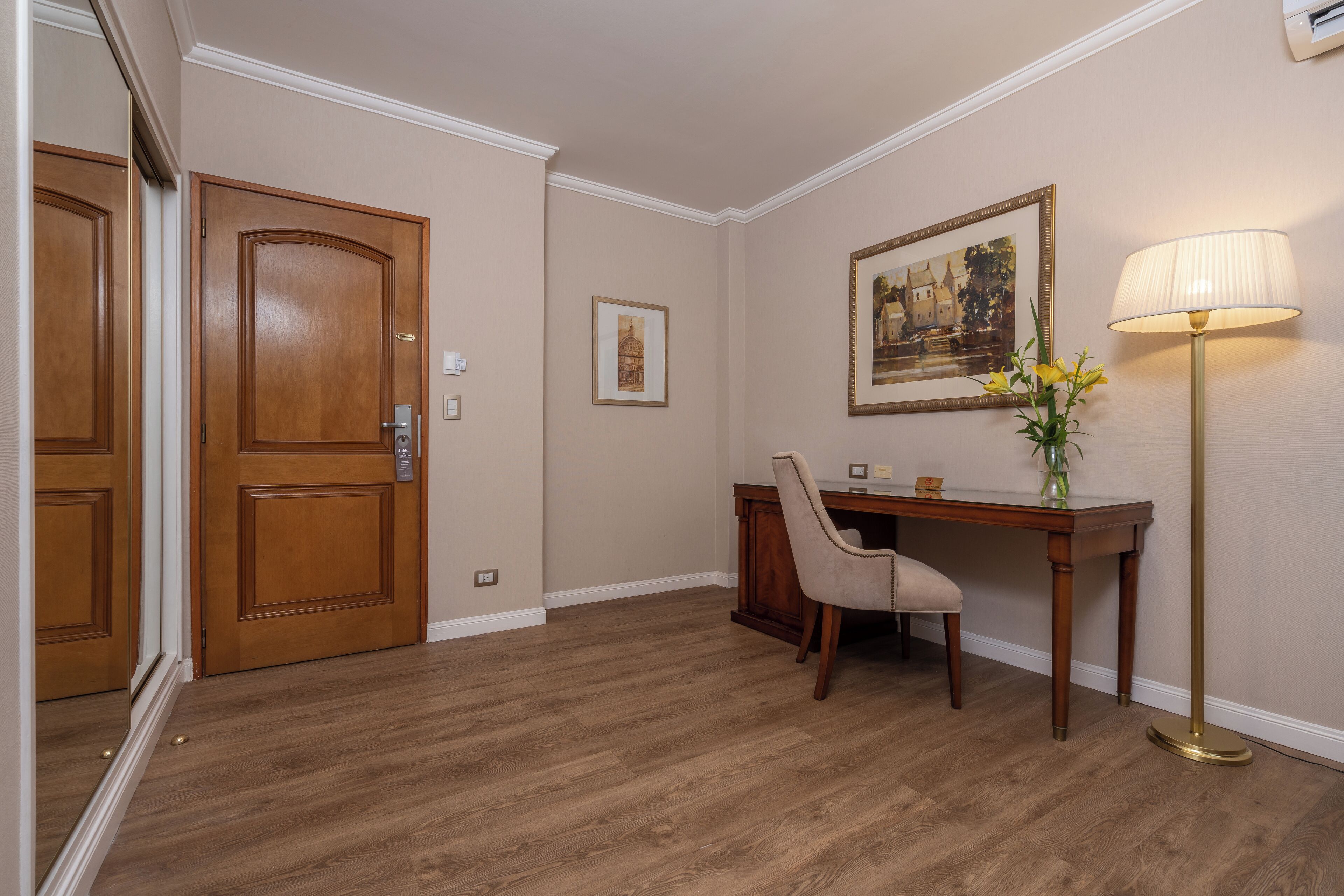 Una habitación con puerta de madera, un escritorio con silla, un cuadro en la pared y una lámpara de pie.