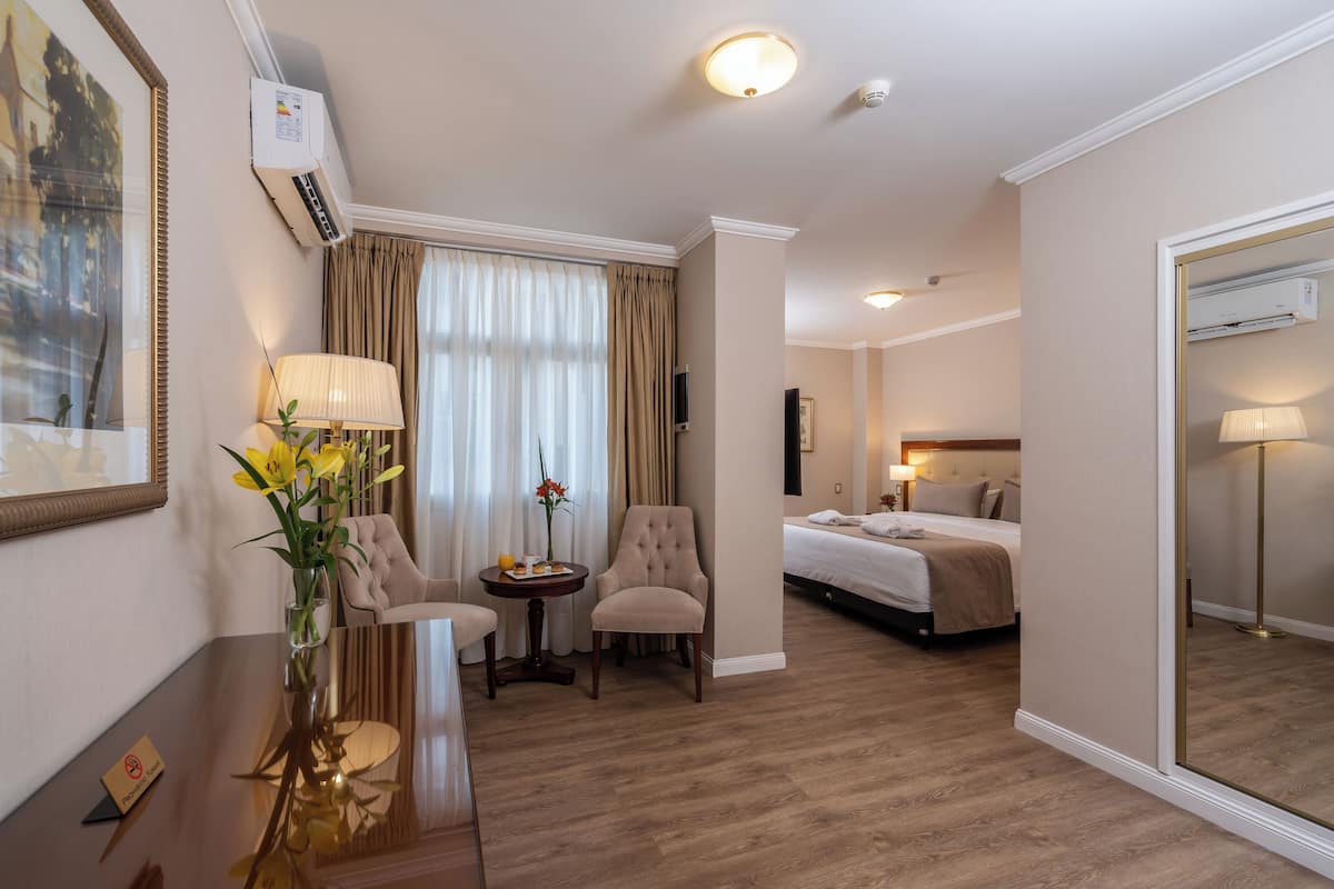 A hotel room with a bed, two armchairs, a small table with a vase of flowers, a mirror, and a wall-mounted air conditioner.