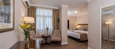A hotel room with a bed, two armchairs, a small table with a vase of flowers, a mirror, and a wall-mounted air conditioner.