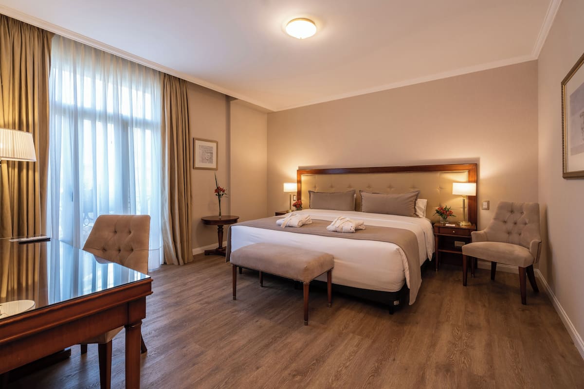 A hotel room with a large bed, a desk, a chair, and a bench.