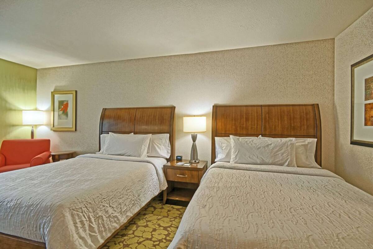 A hotel room with two beds, a nightstand, a lamp, a framed picture, and a red chair.