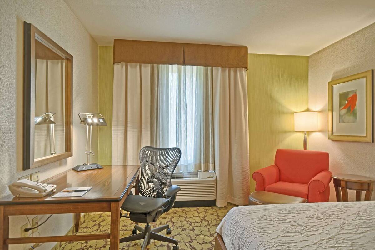 A hotel room with a bed, a desk with a telephone, a chair, a small table, a lamp, and a window with curtains.