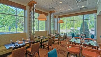 A restaurant with multiple tables and chairs, large windows with blinds, and hanging pendant lights.
