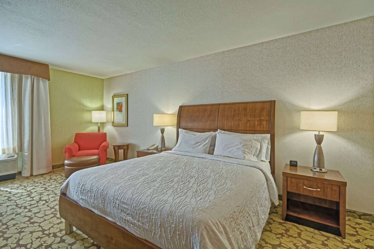 A hotel room with a large bed, two bedside tables with lamps, a red armchair, and a window with curtains.