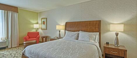 A hotel room with a large bed, two bedside tables with lamps, a red armchair, and a window with curtains.