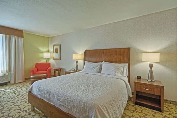 A hotel room with a large bed, two bedside tables with lamps, a red armchair, and a window with curtains.