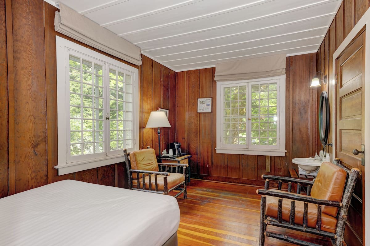 A wooden room with a bed, two chairs, a desk with a lamp, and a sink.