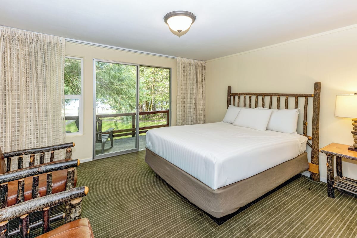 A hotel room with a large bed, a wooden headboard, a small table with a lamp, and a view of a green outdoor area through a sliding glass door.