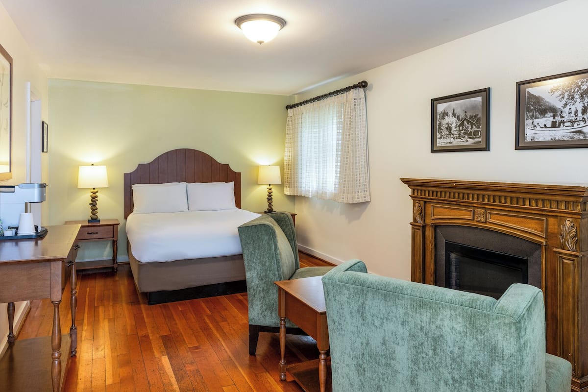 A hotel room with a bed, two bedside lamps, a desk with a coffee maker, a chair, a fireplace, and two framed pictures on the wall.