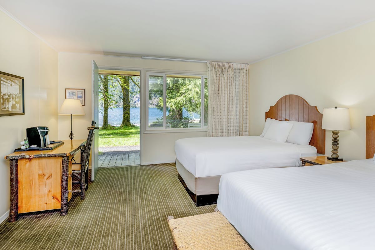 A hotel room with two beds, a wooden headboard, a desk with a lamp, a view of trees and water outside, and a framed picture on the wall.