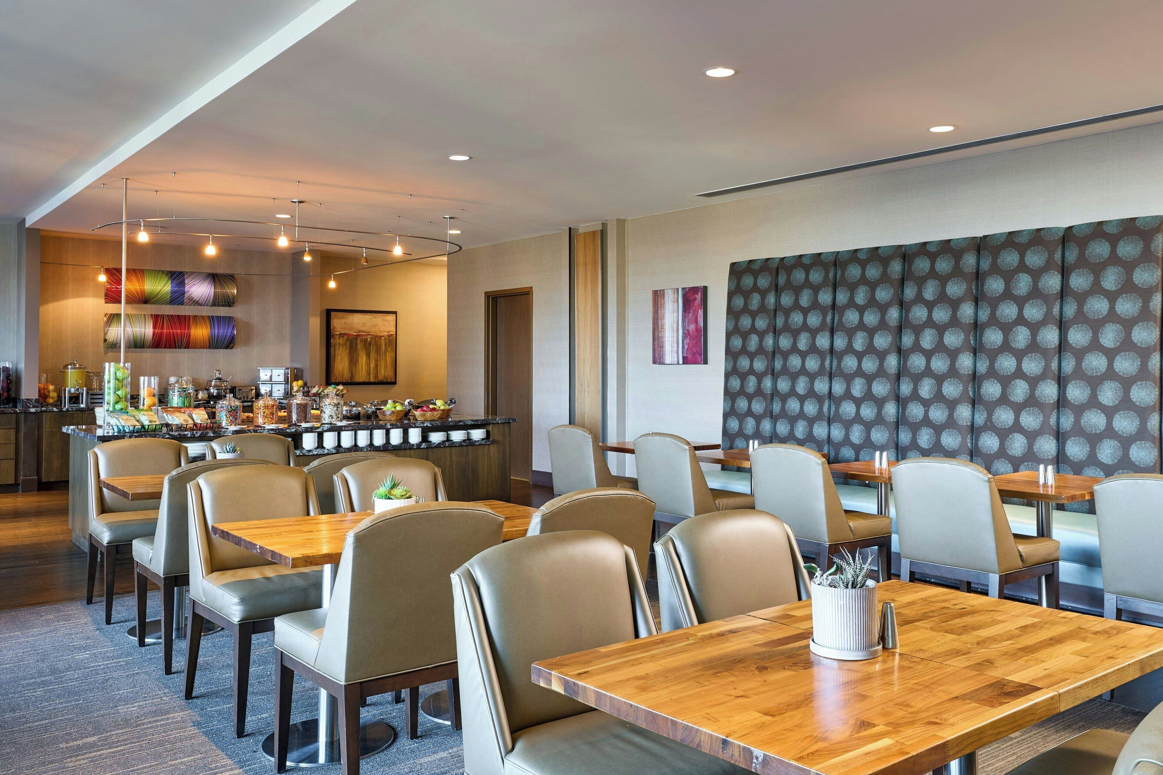 A modern dining area with wooden tables and cushioned chairs, a bar with colorful decor, and a patterned wall.