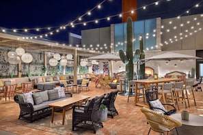 Un espacio al aire libre con mobiliario de mimbre, luces colgantes y un cactus como centro de atención.