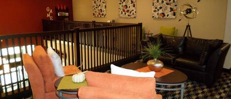 A cozy lounge area with a black leather sofa, orange chairs, a small round table with a plant, and a decorative wall with floral patterns.