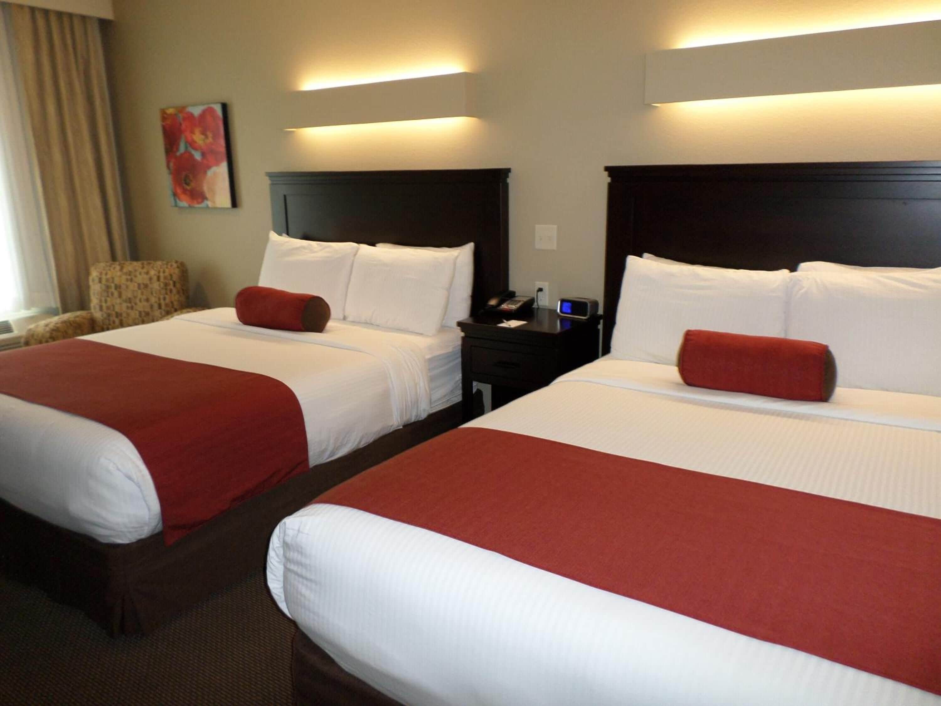A hotel room with two beds, each with white linens and red accents, a nightstand with a phone and a lamp, and a chair.