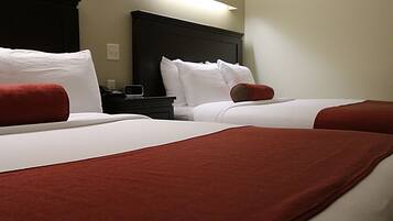 Two beds with white linens and red accents, a dark headboard, and a small bedside table with a lamp.