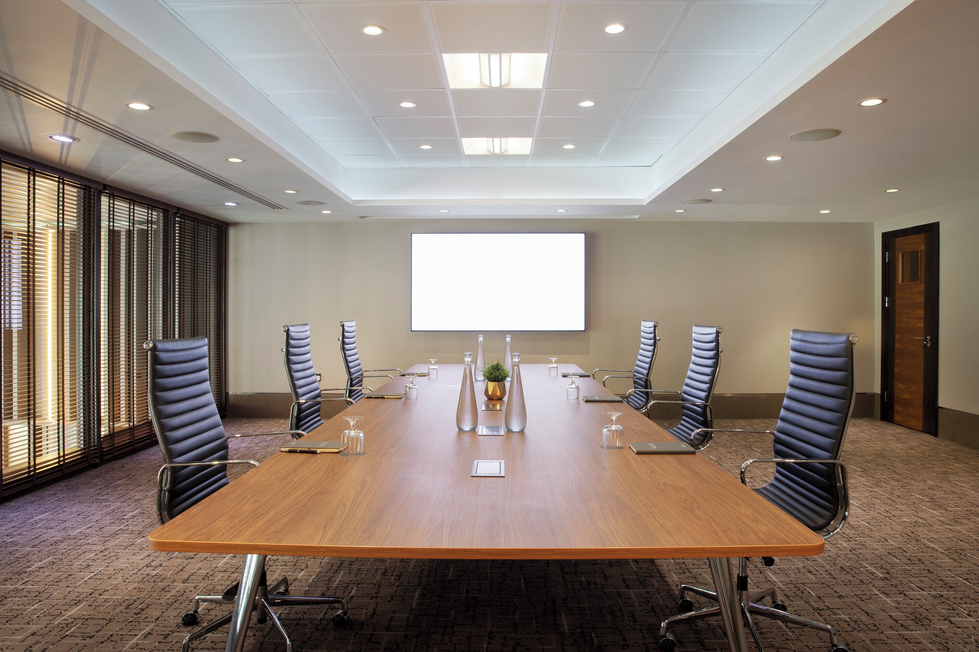 A conference room with a long table, six chairs, a projector screen, and a large window with blinds.