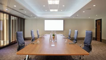 Een vergaderzaal met een lange tafel, zes stoelen, een projectorscherm en een groot raam met jaloezieën.
