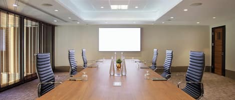 A conference room with a long table, six chairs, a projector screen, and a large window with blinds.
