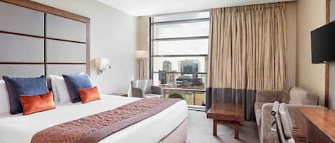 A hotel room with a large bed, a desk, a chair, and a view of the city through a large window.