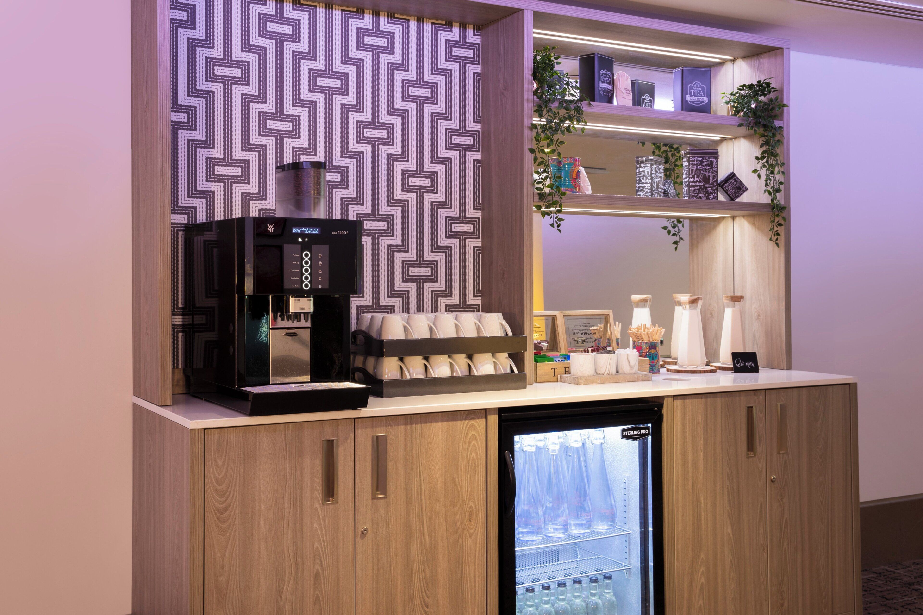 A modern hotel reception area with a coffee machine, a mini-fridge, and a display of cups and saucers.