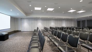 Un salón de conferencias con filas de sillas negras, una pantalla de proyector y suelo alfombrado.