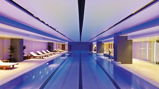 A modern indoor swimming pool with lounge chairs and a glass wall on one side.
